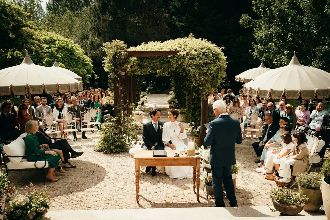 Buitenceremonie in een tuin met een klimopboog en witte parasols